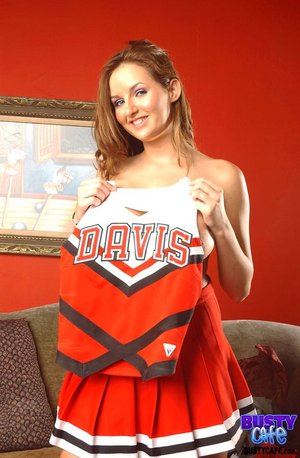 Busty brunette cheerleader takes off her uniform and exposes her large breasts while standing in the red living room